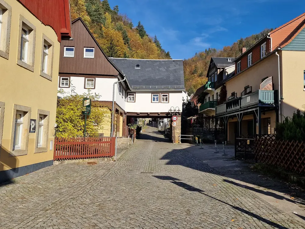 Dorfstraße in Schmilka: Kopfsteinpflaster zwischen Fachwerk- und Umgebindehäusern, dahinter herbstlich bewaldete Felshänge