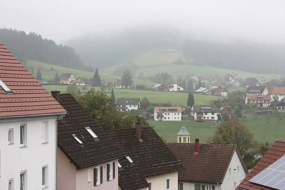 Ortsansicht von Baiersbronn: Häuser mit roten Dächern am Hang, dahinter Wiesen und nebelverhangene Schwarzwaldhöhen