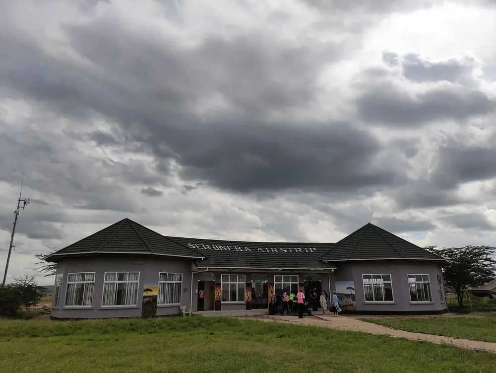 Seronera Airstrip: Abfertigungsgebäude mit Schriftzug unter dunklen Wolken, davor Rasen