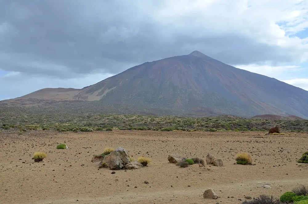 Blick von La Fortaleza über die karge Ebene mit Ginsterbüschen zum Pico del Teide unter grauen Wolken