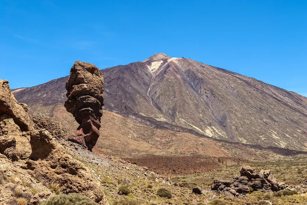 Roque Cinchado: schmaler, verwitterter Felsturm vor dem mächtigen Kegel des Pico del Teide unter blauem Himmel