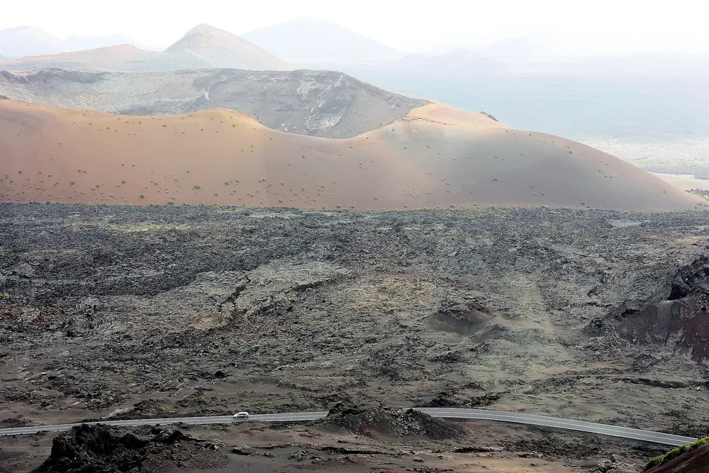 Blick über ein dunkles Lavafeld zur hellen, weiten Caldera del Corazoncillo, am Rand die Straße LZ-67