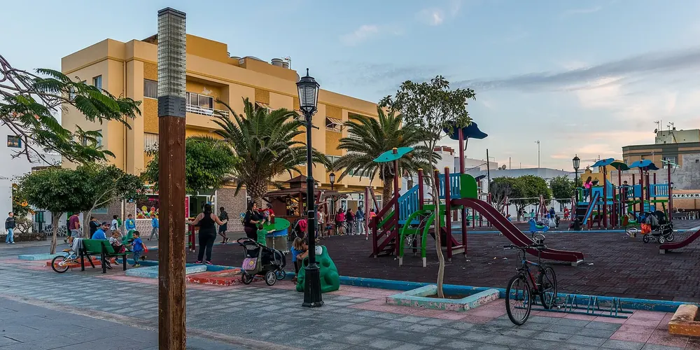 Platz in Corralejo mit Spielplatz, Palmen und Laternen vor farbigen Wohnhäusern im Abendlicht