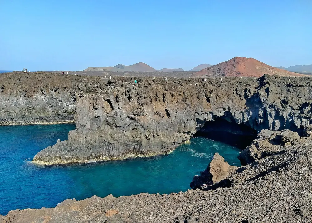 Los Hervideros: schroffe Lavaklippen mit Höhlen über tiefblauem Meer, dahinter rote Vulkankegel
