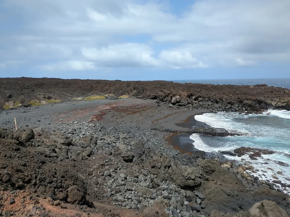 Playa de la Madera: dunkle Lavaküste mit kleiner Sandbucht und weißer Brandung unter bewölktem Himmel
