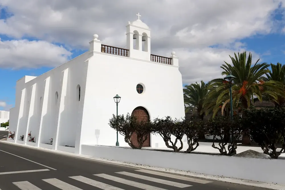 Iglesia San Isidro Labrador in Uga: weiße Kirche mit Glockengiebel und Holzportal zwischen Palmen