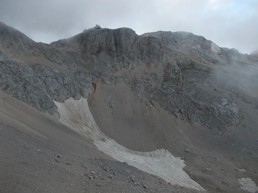 Triglavski dom na Kredarici: Berghütte auf dem kahlen Grat unter grauen Felswänden, am Hang Schneereste