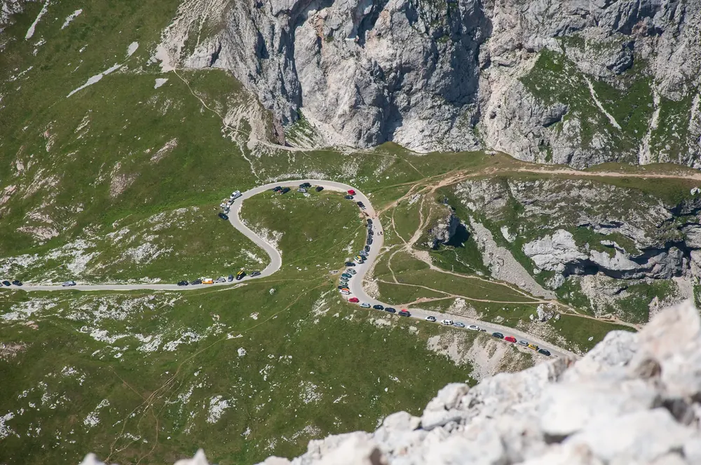 Serpentinen der Mangart-Straße auf grünen Almhängen unter den Kalkwänden des Mangart, gesäumt von Autos