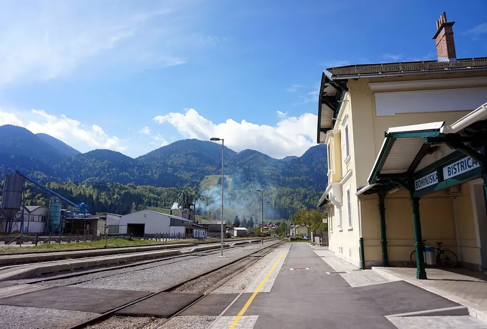 Bahnhof Bohinjska Bistrica: gelbes Empfangsgebäude mit Bahnsteigdach und Gleisen vor bewaldeten Bergen