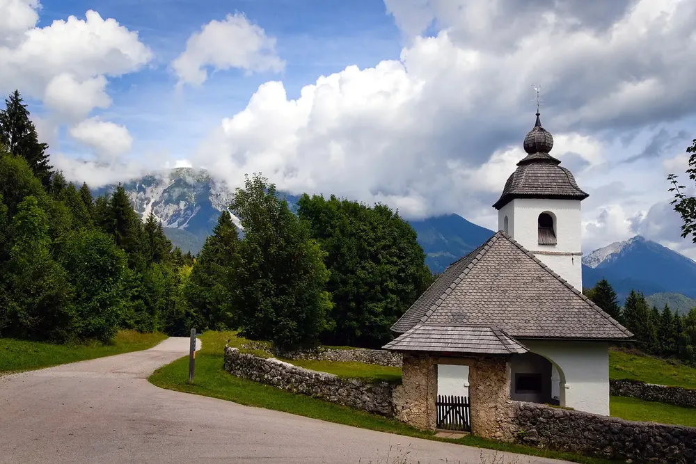 Kirche St. Katharina bei Zasip: weiße Kapelle mit Zwiebelturm an einem Weg, dahinter Wald und Berge