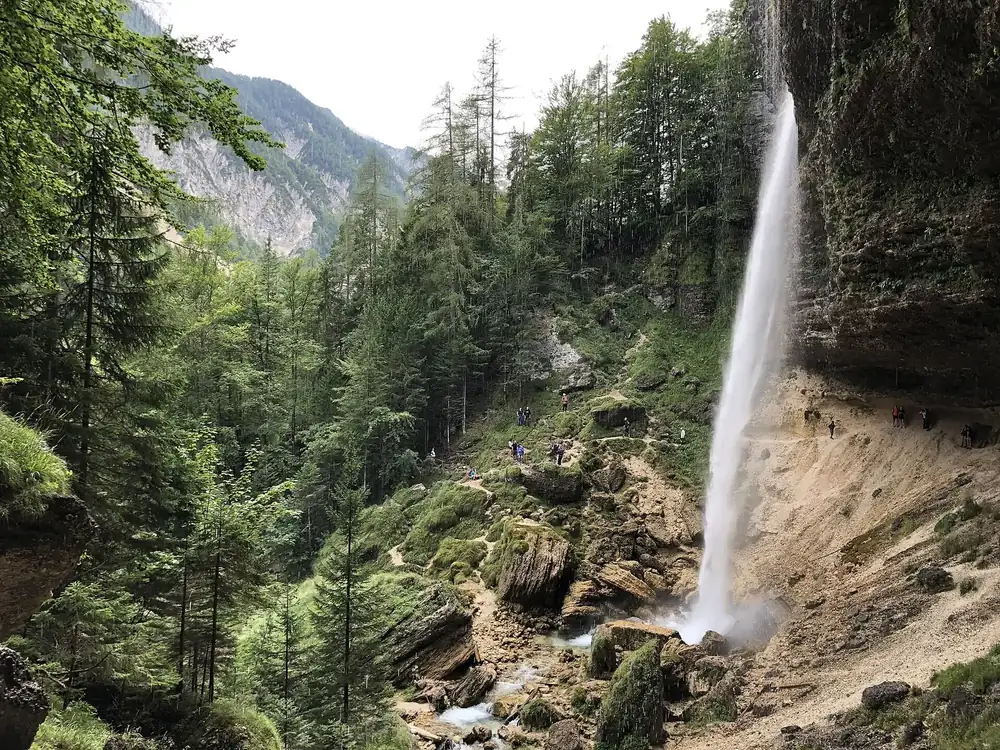 Peričnik-Wasserfall: Wasser stürzt über eine überhängende Felskante in den Wald, dahinter Berghänge