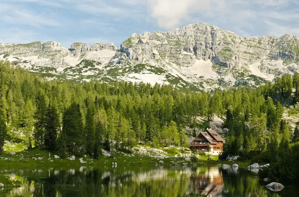 Koča pri Triglavskih jezerih: Berghütte am Ufer eines Bergsees, dahinter Nadelwald und helle Kalkgipfel