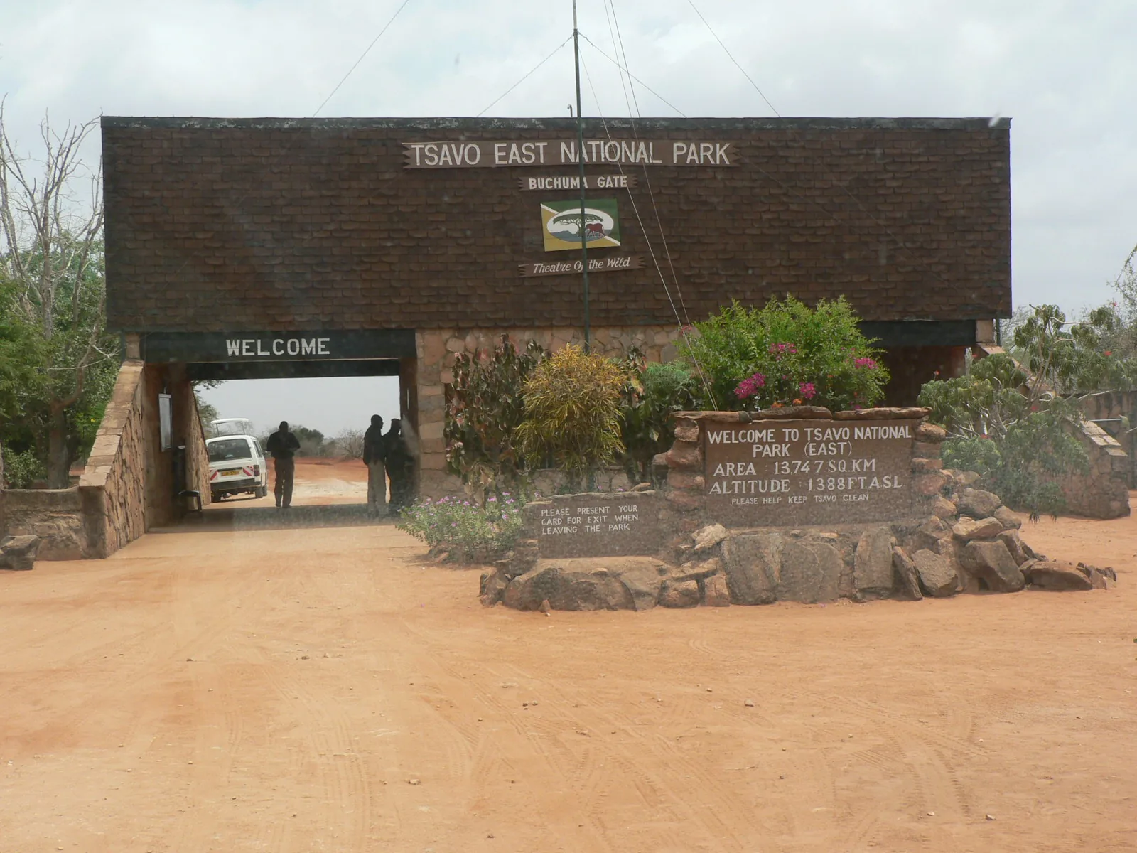 Das Bachuma Gate von Tsavo East mit Torbogen, Willkommensschild und Steinmauer auf roter Erde