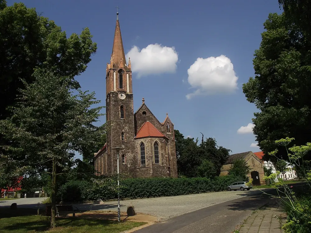 Backsteinkirche in Hohensaaten mit spitzem Turm und Uhr, davor eine Dorfstraße und Bäume unter blauem Himmel