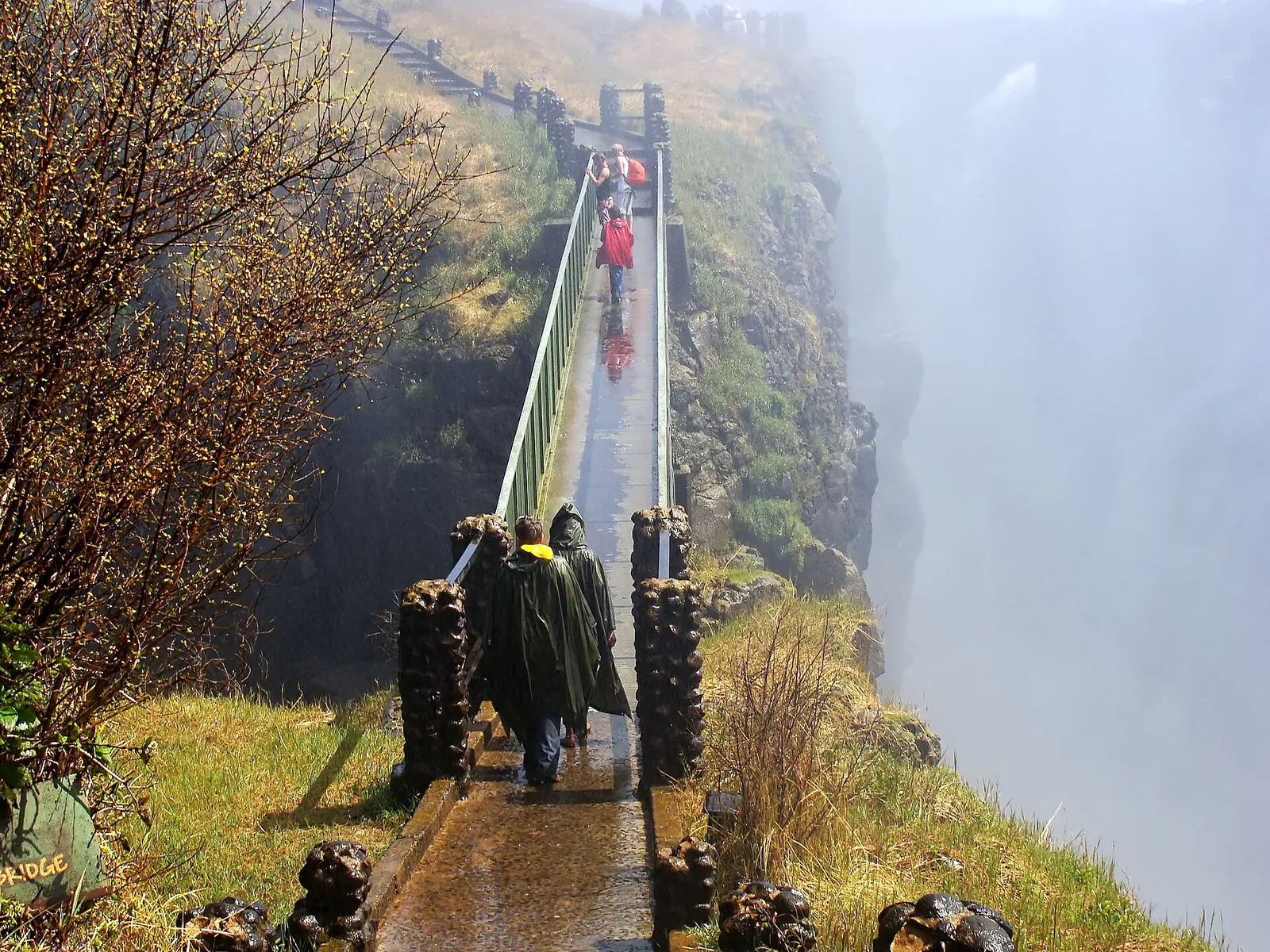 Die Knife Edge Bridge auf der sambischen Seite der Victoriafälle: eine schmale Fußgängerbrücke im Sprühnebel, Besucher in Regencapes