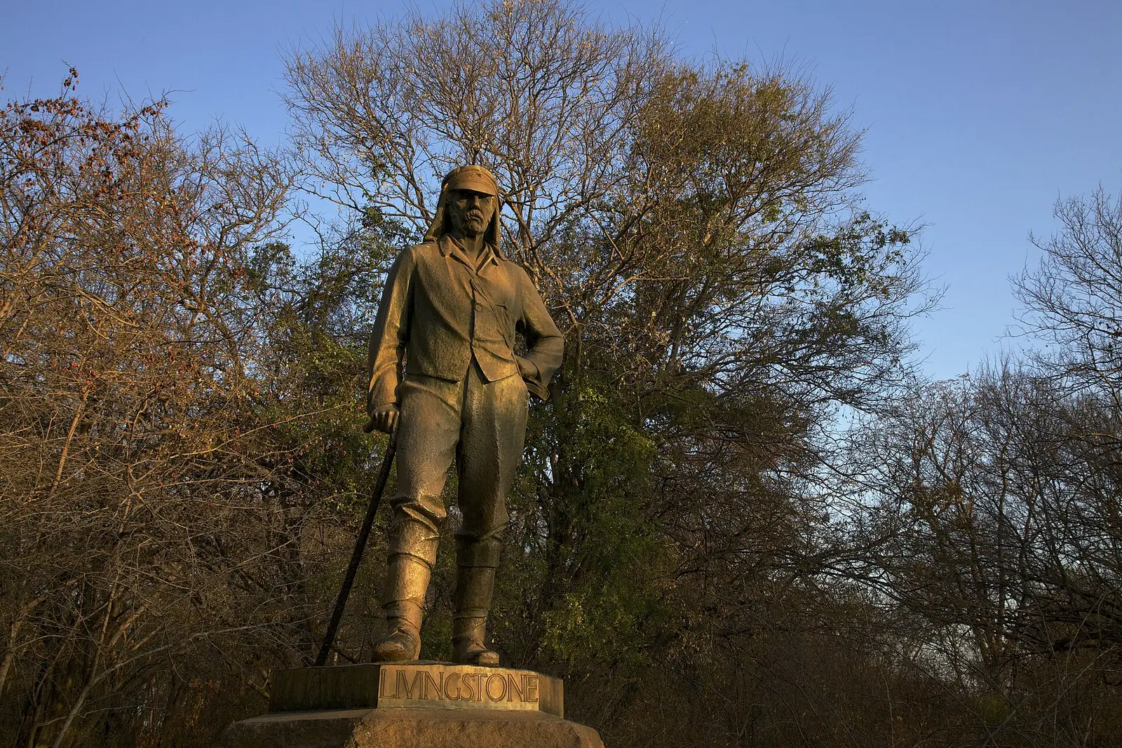 Bronzestatue von David Livingstone an den Victoriafällen in Simbabwe, im warmen Morgenlicht vor kahlen Bäumen