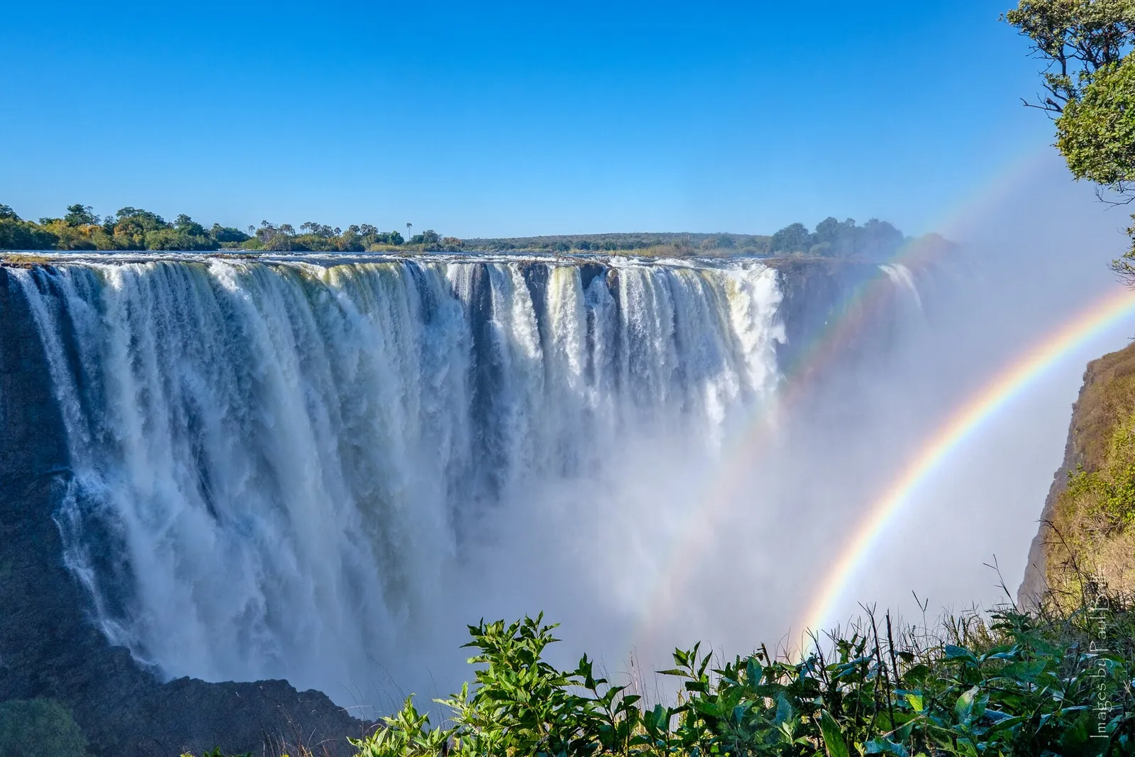 Die Main Falls der Victoriafälle von der simbabwischen Seite: eine breite Wasserwand über der Schluchtkante, davor ein doppelter Regenbogen in der Gischt