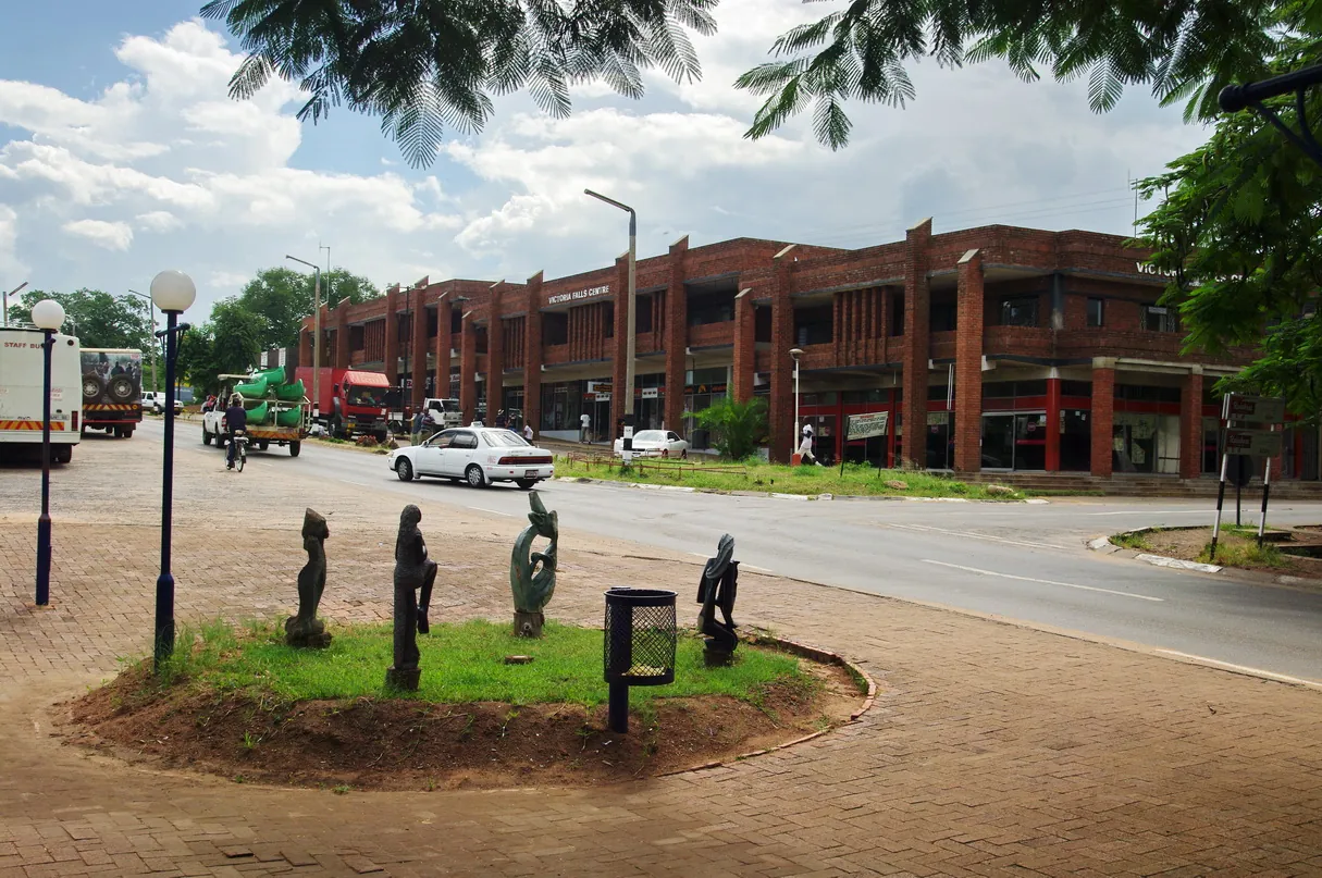 Straße im Ort Victoria Falls mit Holzskulpturen auf einer Verkehrsinsel und einem roten Backstein-Geschäftshaus