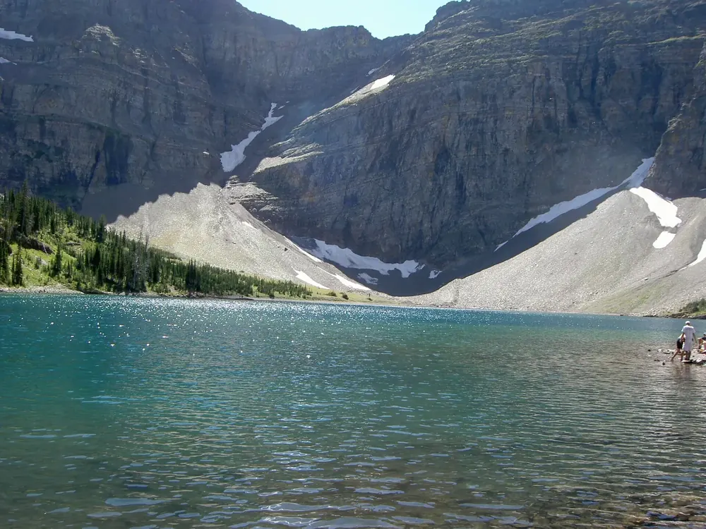 Crypt Lake: türkisfarbener Bergsee in einem Felskessel mit Geröllhängen, Schneeresten und steilen Wänden