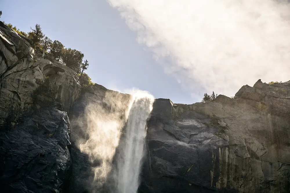 Bridalveil Fall: Wasserfall stürzt über eine dunkle Granitkante, der Sprühnebel leuchtet vor hellem Himmel