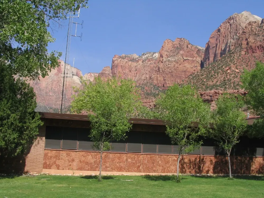 Zion Human History Museum: flacher Backsteinbau zwischen jungen Bäumen, dahinter rote Felswände des Zion Canyon