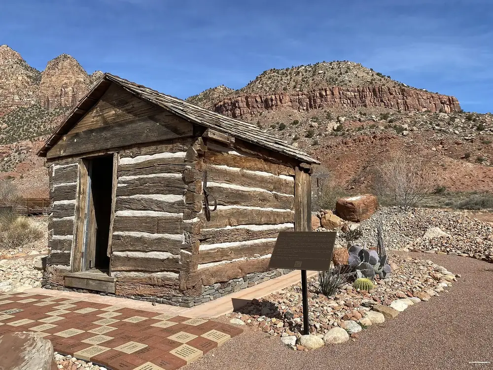 Historischer Zehntspeicher aus Holzbalken in Springdale, dahinter rote Felswände am Eingang zum Zion Canyon