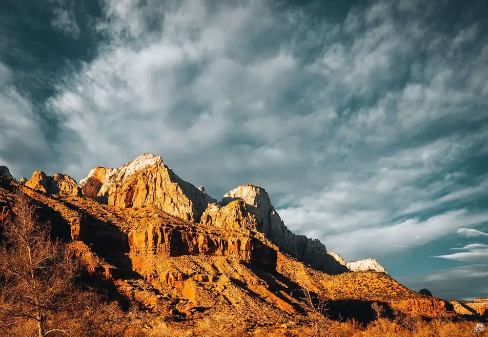 The Watchman im goldenen Abendlicht: zerklüfteter Sandsteingipfel über rötlichen Felsstufen und dramatischem Wolkenhimmel