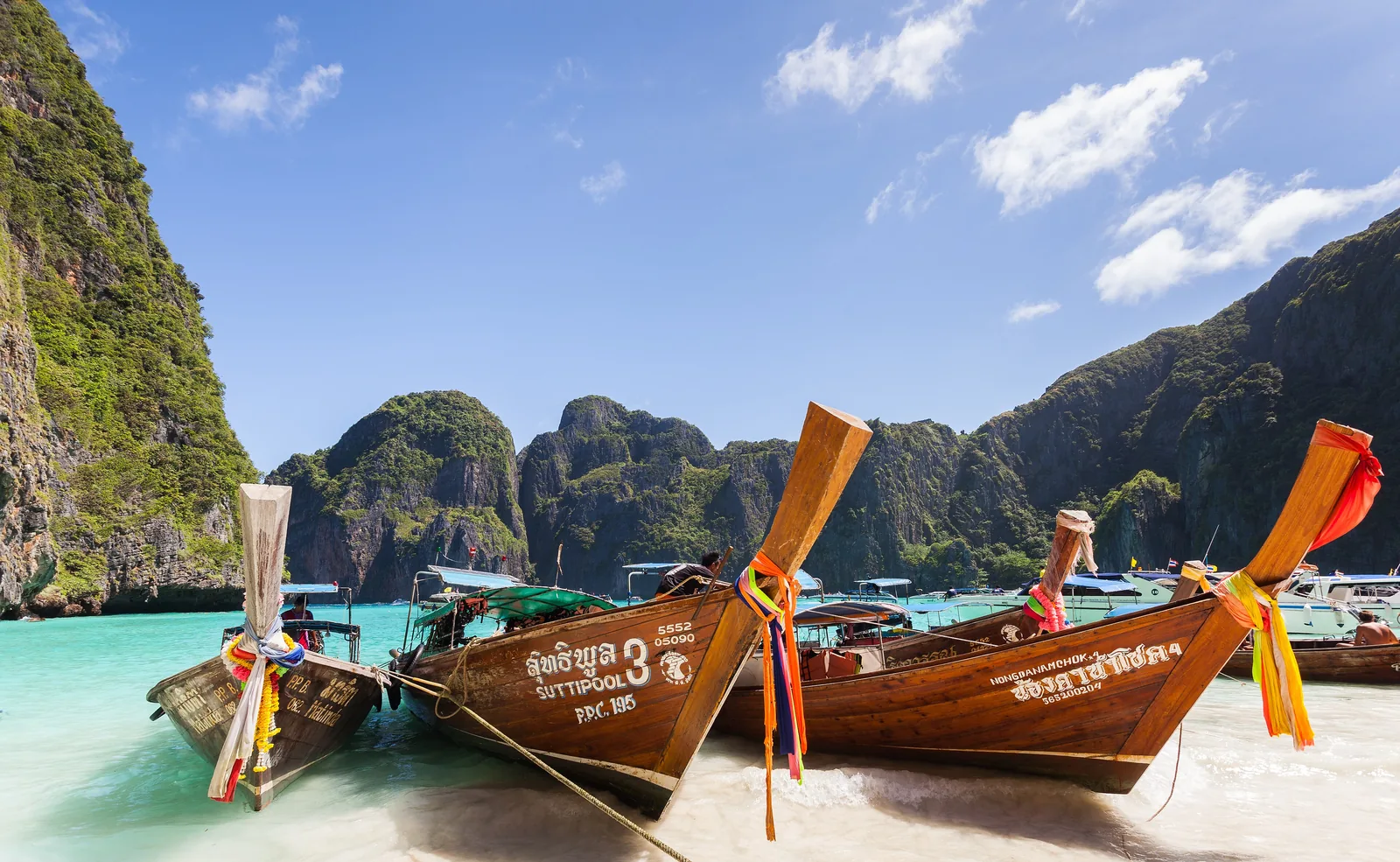 Bunt geschmückte Langheckboote liegen am hellen Strand der Maya Bay, dahinter die Kalkfelsen der Bucht