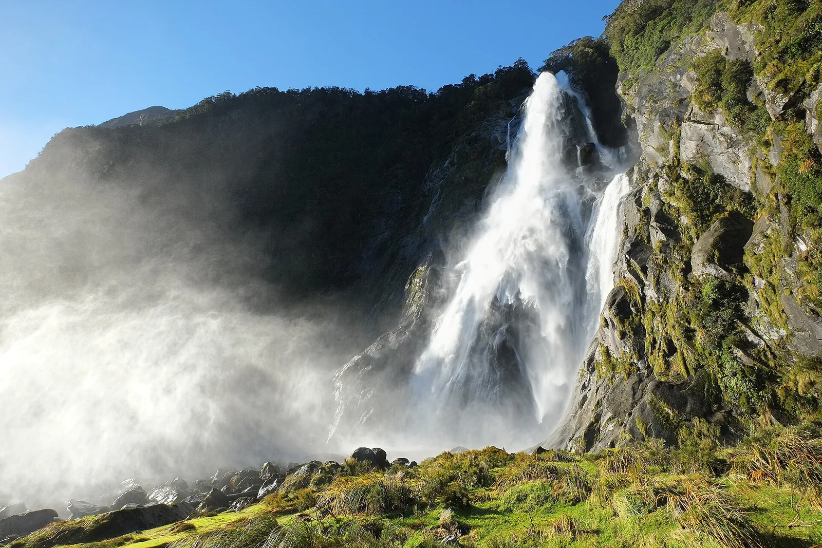 Die Bowen Falls am Milford Sound stürzen in der Sonne über eine Felskante, die Gischt steigt als weiße Wolke auf