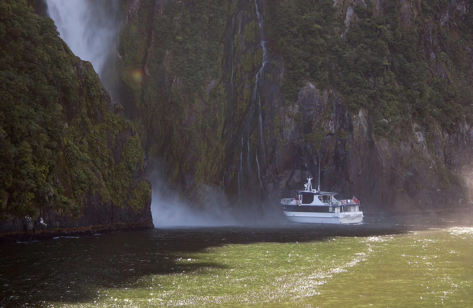 Ein Ausflugsschiff auf dem Milford Sound fährt an die Gischt der Stirling Falls unter einer steilen, bewaldeten Felswand heran