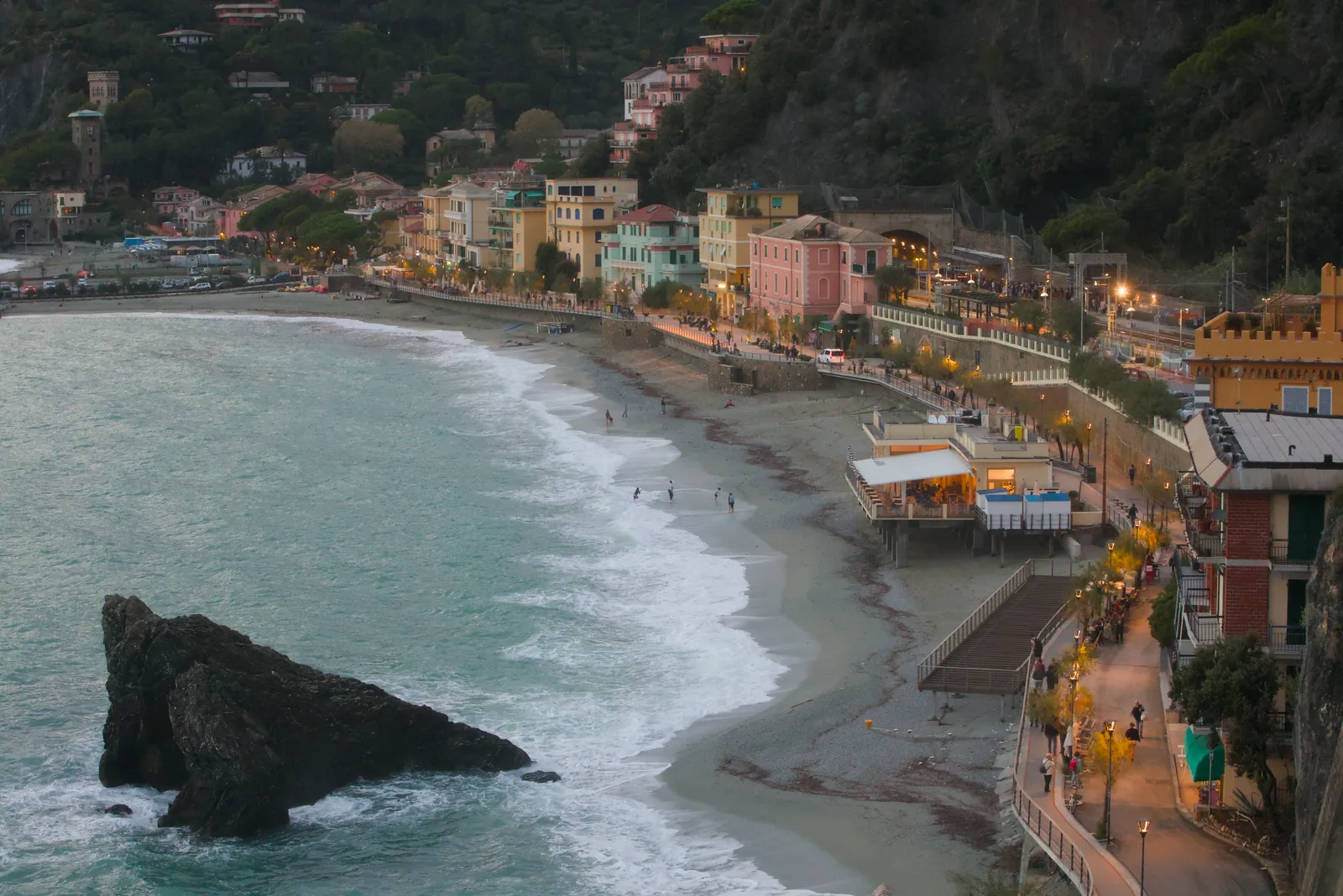 Monterosso al Mare in der Abenddämmerung: der Strand von Fegina mit einem Felsen in der Brandung, dahinter die beleuchtete Uferpromenade