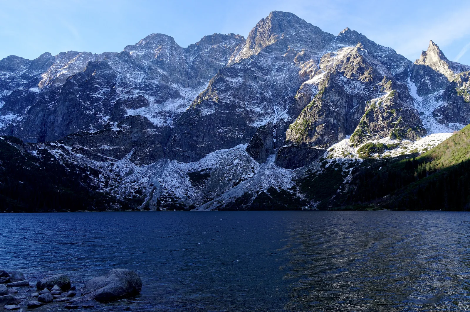Der Morskie Oko im Tatra-Nationalpark im Oktober, dahinter die verschneiten Mięguszowieckie Szczyty, im Vordergrund Steine am Ufer