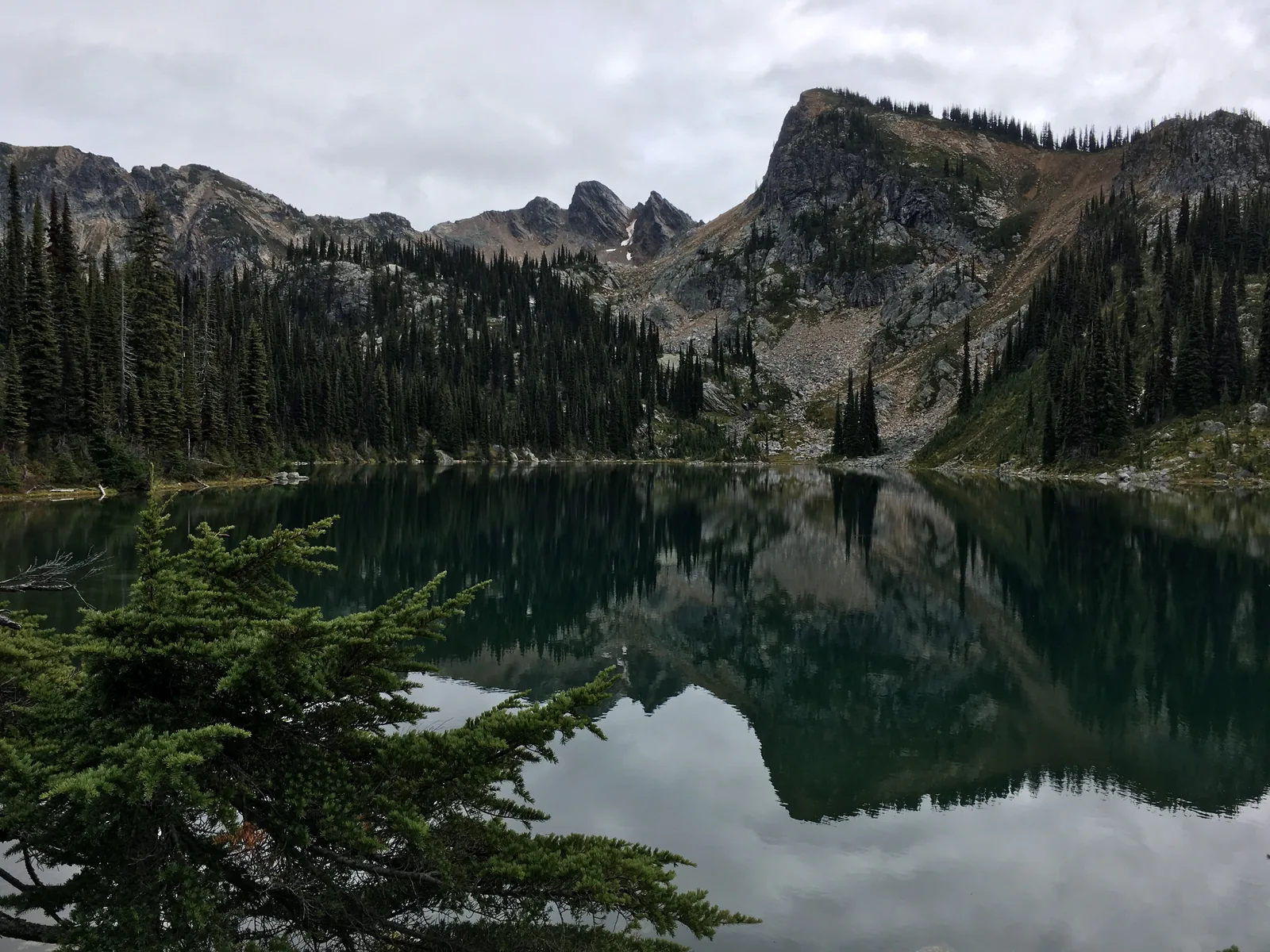 Der stille Eva Lake spiegelt eine gezackte, teils schneebedeckte Felswand; am Ufer stehen einzelne Nadelbäume