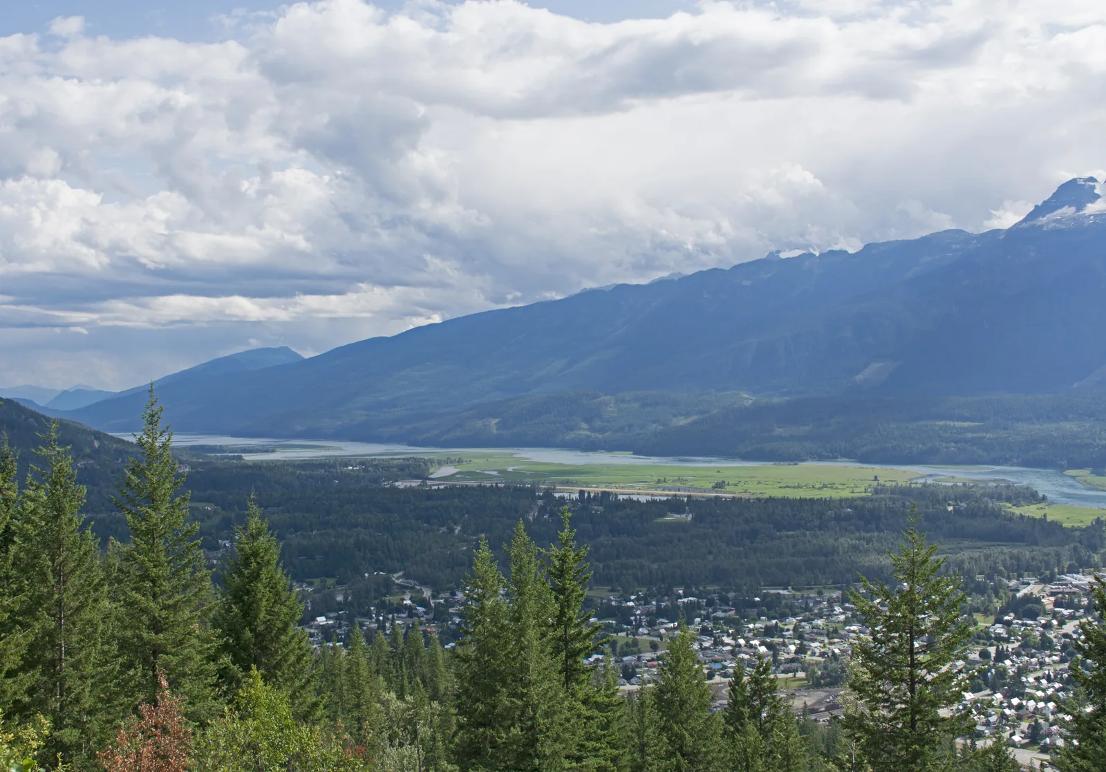 Blick von der Bergstraße über bewaldete Hänge auf die Stadt Revelstoke und das breite, silbrige Band des Columbia River, dahinter Bergketten