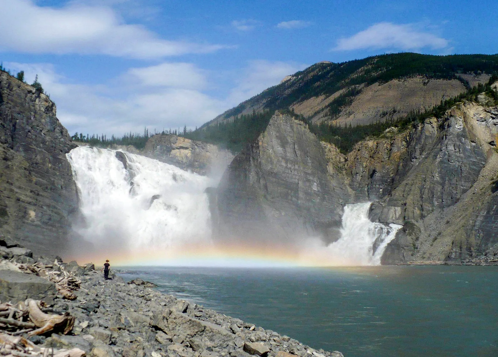 Náįlįcho (Virginia Falls) vom Ufer aus: zwei mächtige Wasserfallarme stürzen in eine Schlucht, in der Gischt steht ein Regenbogen, am Ufer eine winzige Gestalt