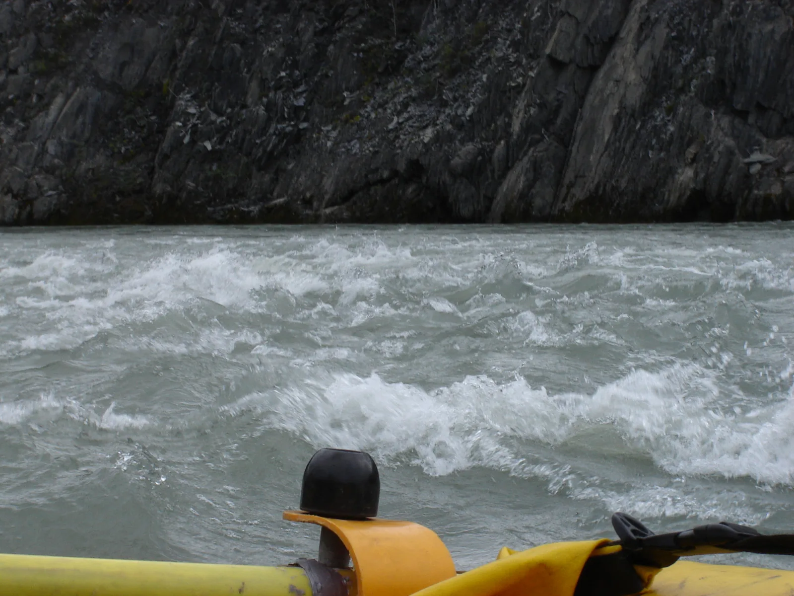 Wildwasser auf dem Nahʔą Dehé aus der Perspektive eines Schlauchboots: schäumende Wellen vor einer dunklen Felswand, im Vordergrund ein Helm