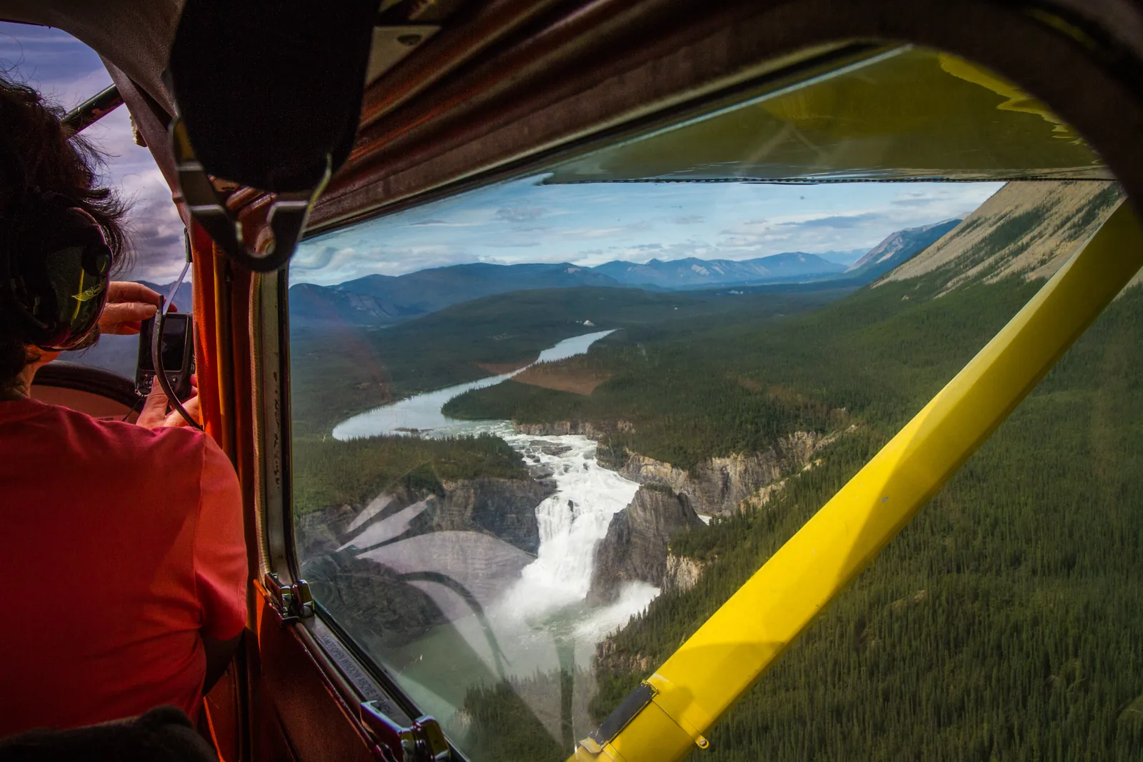 Blick aus dem Fenster eines kleinen Flugzeugs auf Náįlįcho (Virginia Falls) und den South Nahanni River, im Vordergrund Pilot und Tragflächenstrebe