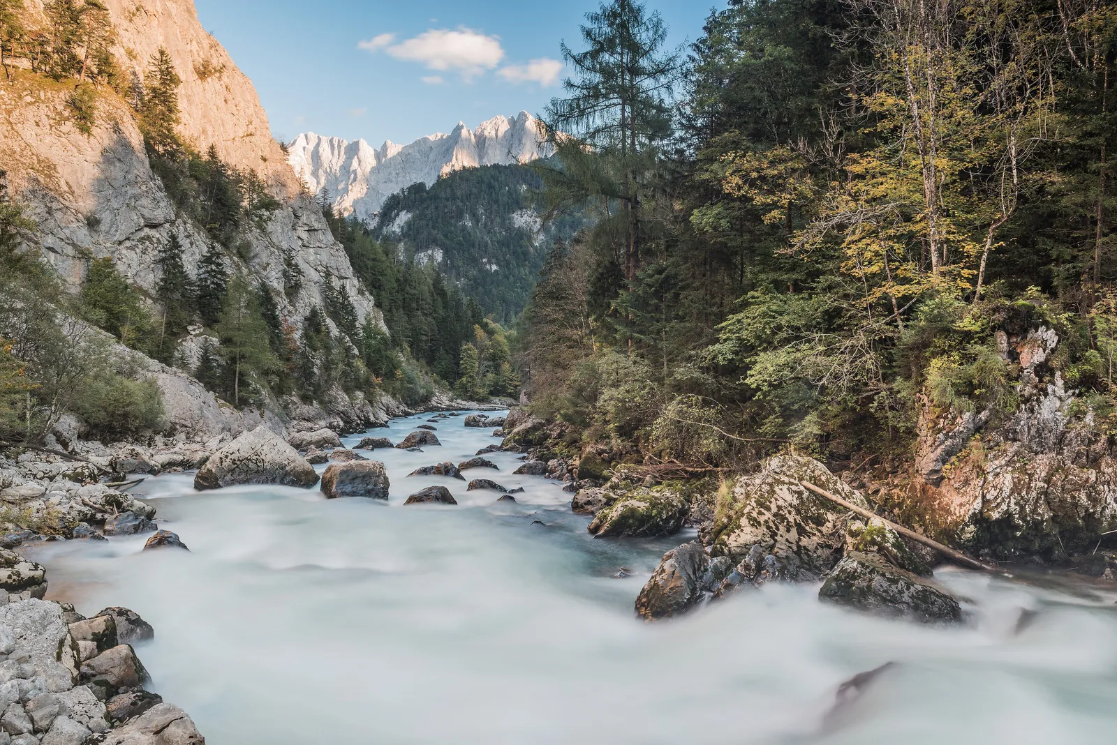 Die Enns schäumt am Gesäuseeingang im Nationalpark Gesäuse zwischen Felsblöcken, links eine sonnige Felswand, hinten verschneite Gipfel