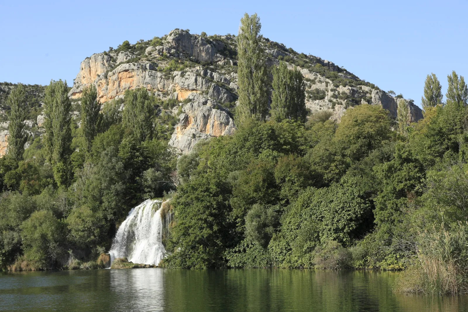 Ein weißer Wasserfall stürzt im Nationalpark Krka zwischen dichtem Grün in einen ruhigen See, dahinter hohe Pappeln und helle Kalkfelsen unter blauem Himmel