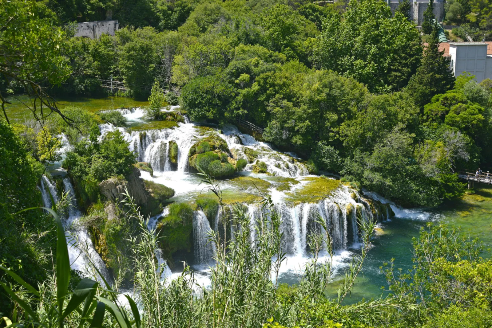 Von oben gesehen stürzt die Krka im Nationalpark Krka über mehrere bemooste Kalktuffstufen mit kleinen bewachsenen Inseln, ringsum dichtes Grün und Holzstege