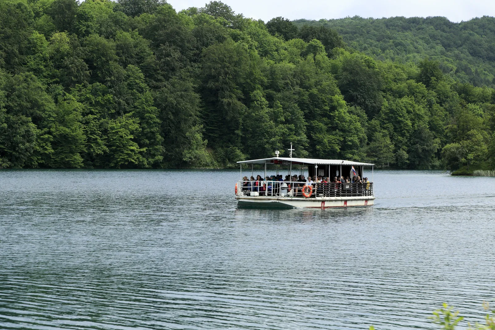 Ein weißes Ausflugsboot voller Fahrgäste gleitet über den grau schimmernden Kozjak-See im Nationalpark Plitvicer Seen vor einem dicht bewaldeten Ufer