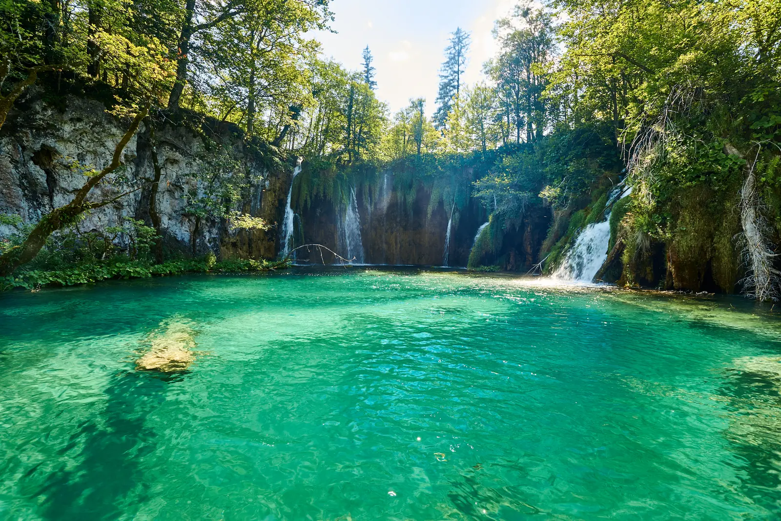 Türkisgrünes, klares Wasser der Oberen Seen im Nationalpark Plitvicer Seen vor einer bewaldeten Tuffkante, über die mehrere weiße Wasserfälle des Galovački buk fallen