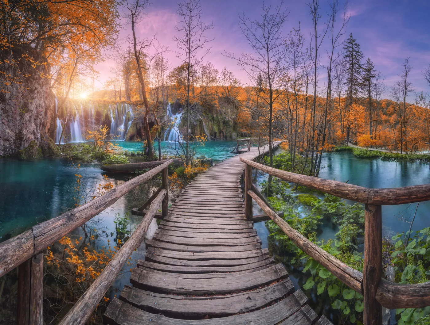 Holzsteg über türkisfarbenem Wasser im Nationalpark Plitvicer Seen, dahinter Wasserfälle und Herbstwald im Abendlicht