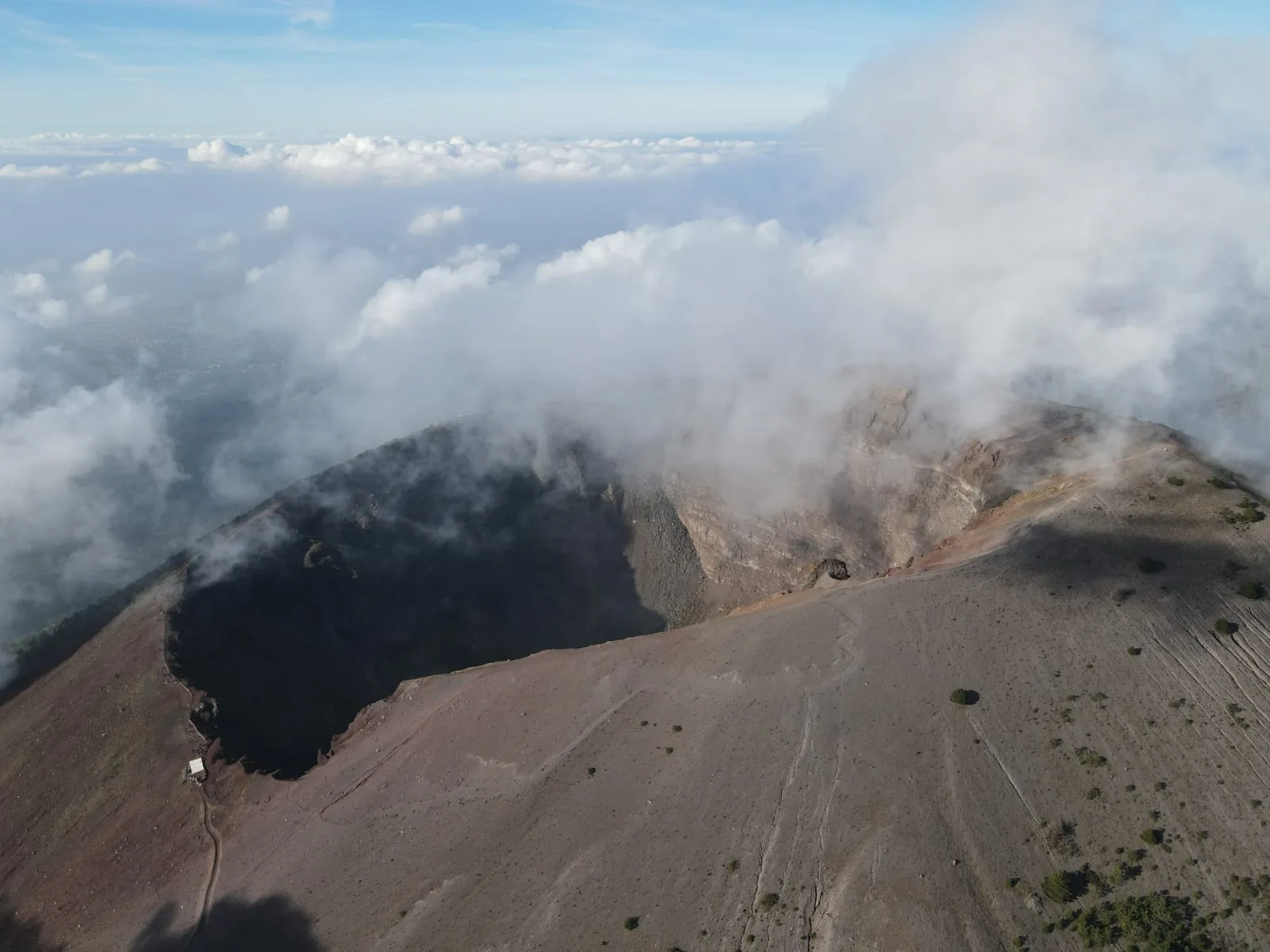 Der Krater des Gran Cono im Nationalpark Vesuv von oben, an seinem Rand ziehen Wolken vorbei