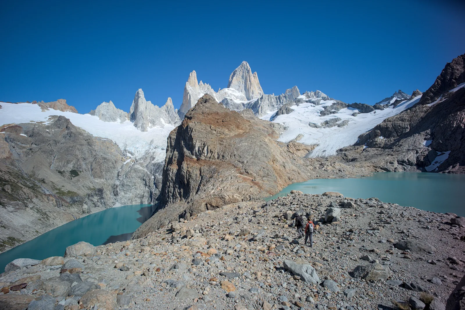 Die Granitnadeln des Fitz Roy über einem Gletscher und zwei türkisfarbenen Lagunen, davor ein Geröllhang mit Wanderern