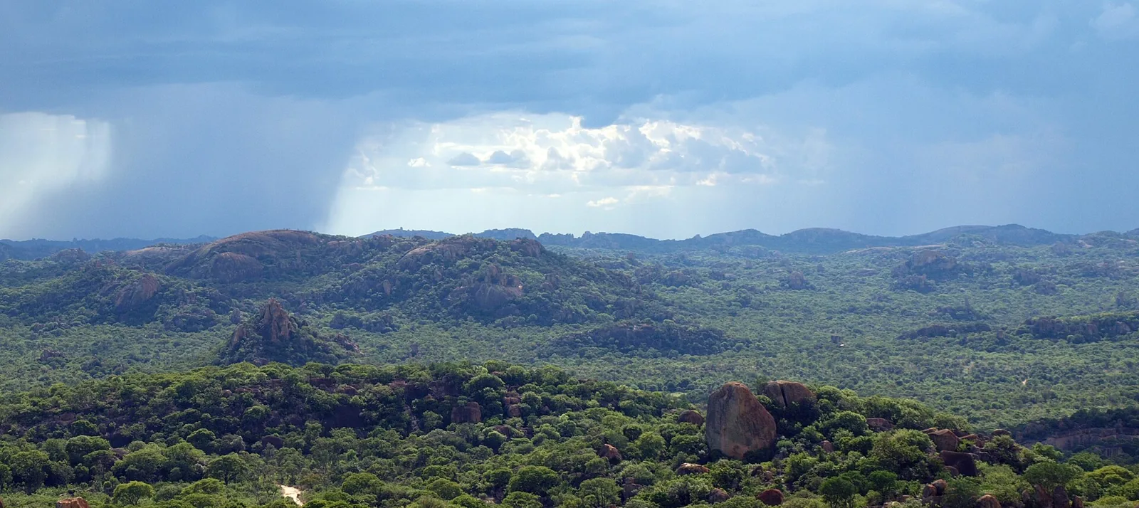 Bewaldete Granithügel und Felskuppen im Matobo-Nationalpark, darüber dunkle Wolken mit einem Regenschauer