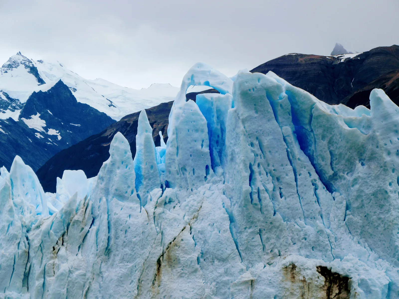 Blaue Eiszacken und ein Bogen in der Front des Perito-Moreno-Gletschers im Nationalpark Los Glaciares, dahinter dunkle Berge
