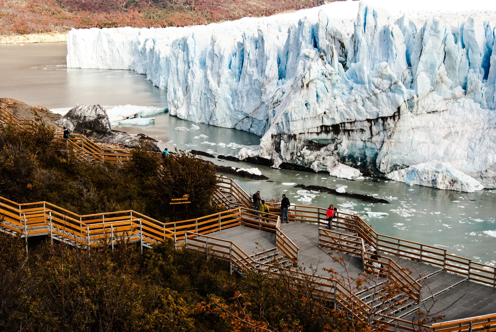 Besucher auf den Holzstegen am Perito-Moreno-Gletscher im Nationalpark Los Glaciares, dahinter die hohe Eisfront und das Wasser des Sees