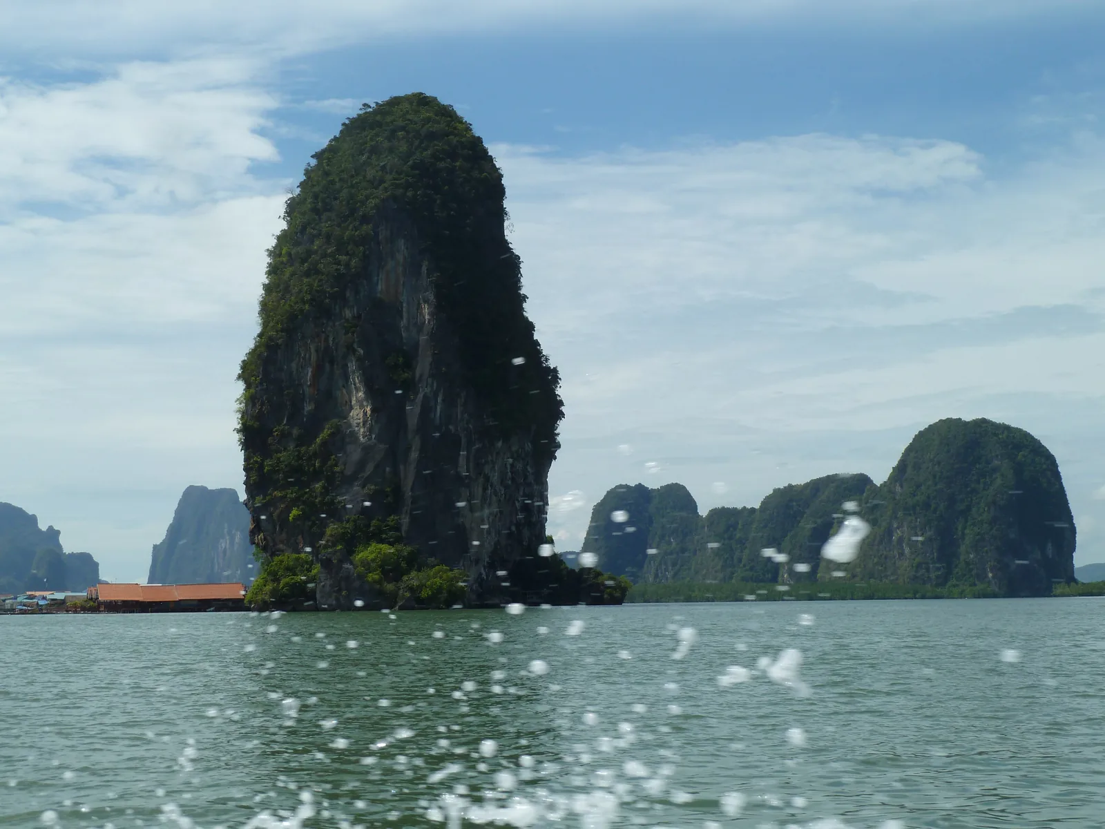 Ein hoher bewachsener Kalkfelsen vor Ko Panyi im Nationalpark Ao Phang Nga, links davor die Dächer des Pfahldorfs am Wasser