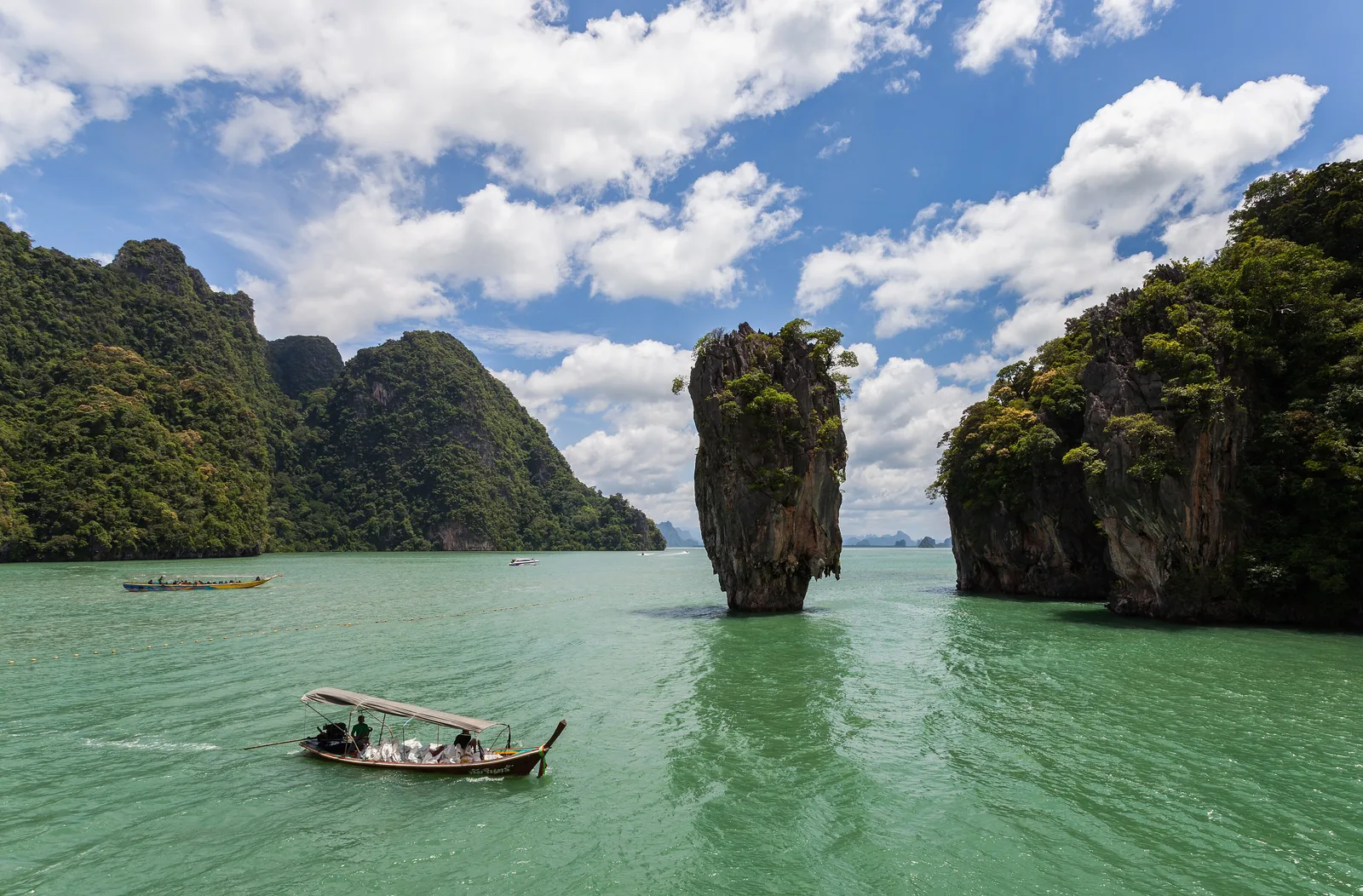 Der schmale Kalkfelsen Ko Tapu ragt im Nationalpark Ao Phang Nga aus grünem Wasser, rechts die Felswand von Khao Phing Kan, vorne ein Langheckboot